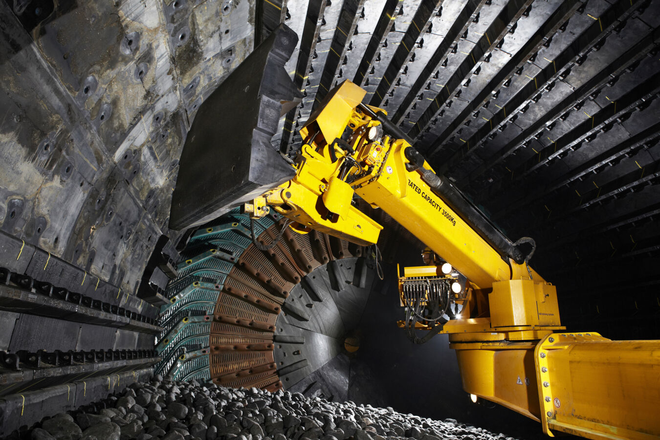 Robot górniczy w tunelu, zbierający kamienie za pomocą dużego chwytaka. W tle widoczna struktura tunelu z metalowymi elementami.