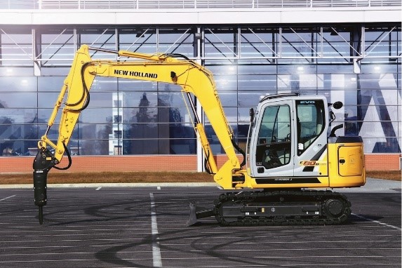 Żółty koparko-ładowarka marki New Holland na tle dużego szklanego budynku.