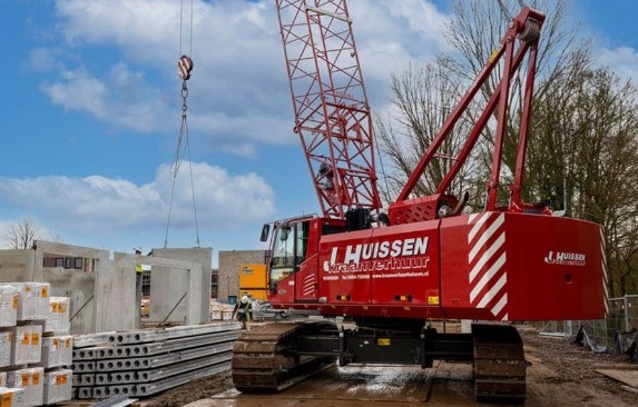 Czerwony dźwig z długim ramieniem na placu budowy, unoszący prefabrykowane elementy betonowe, w otoczeniu materiałów budowlanych i zieleni.
