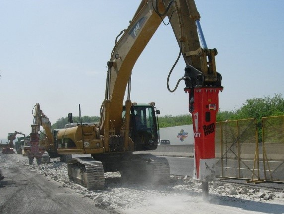Maszyna budowlana z młotem hydraulicznym rozbija nawierzchnię drogi na placu budowy, wokół widoczne są inne maszyny i zieleń w tle.